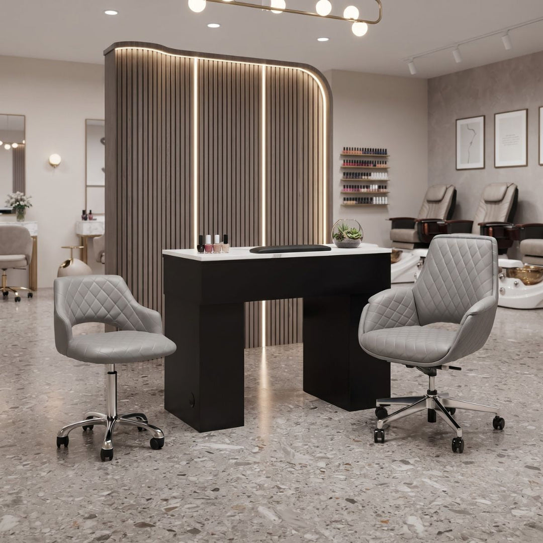 Manicure table and gray quilted chairs in a modern salon; pedicure stations and slatted wall in background.